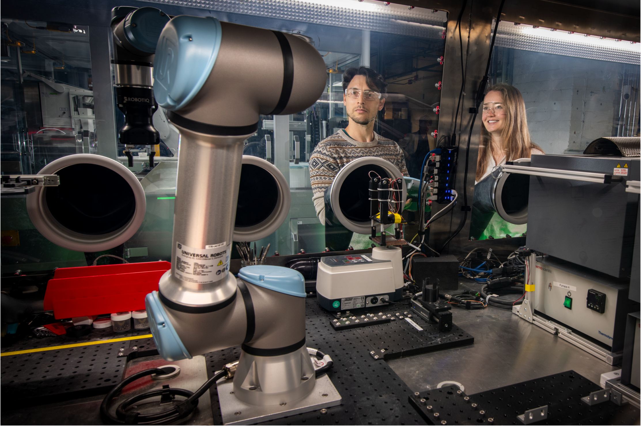 Two researchers wearing safety glasses observe an automated robotic system operating inside a laboratory equipped with enclosed instrumentation and advanced scientific equipment