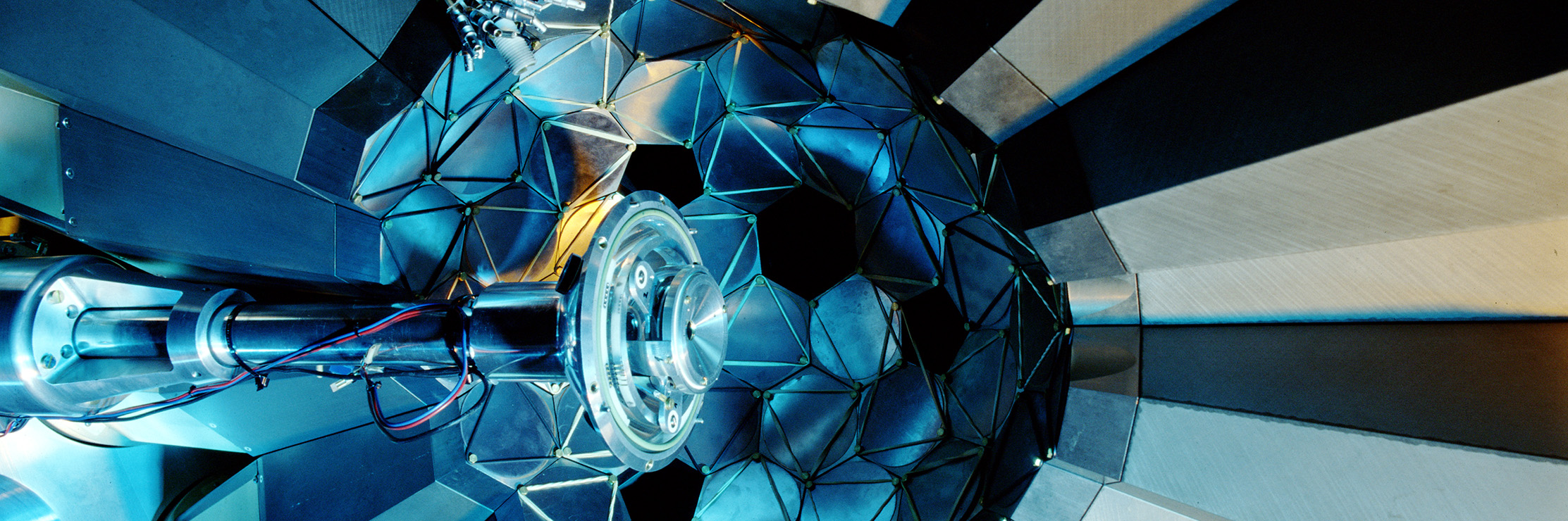 Close-up view inside a scientific instrument, showing a central metal component surrounded by reflective geometric panels illuminated in blue light
