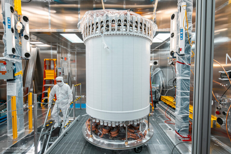 A researcher in protective gear works near a tall, cylindrical scientific instrument with visible wiring. The walls surrounding it are reflective Teflon.