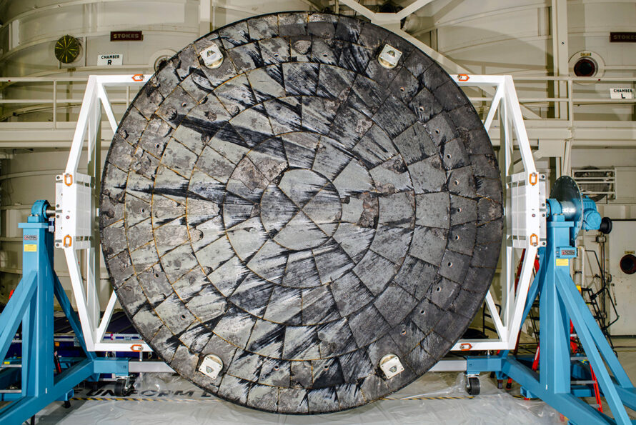 A large circular heat shield with a tiled pattern that shows dark burn marks from the center, mounted on a metal frame in a lab.