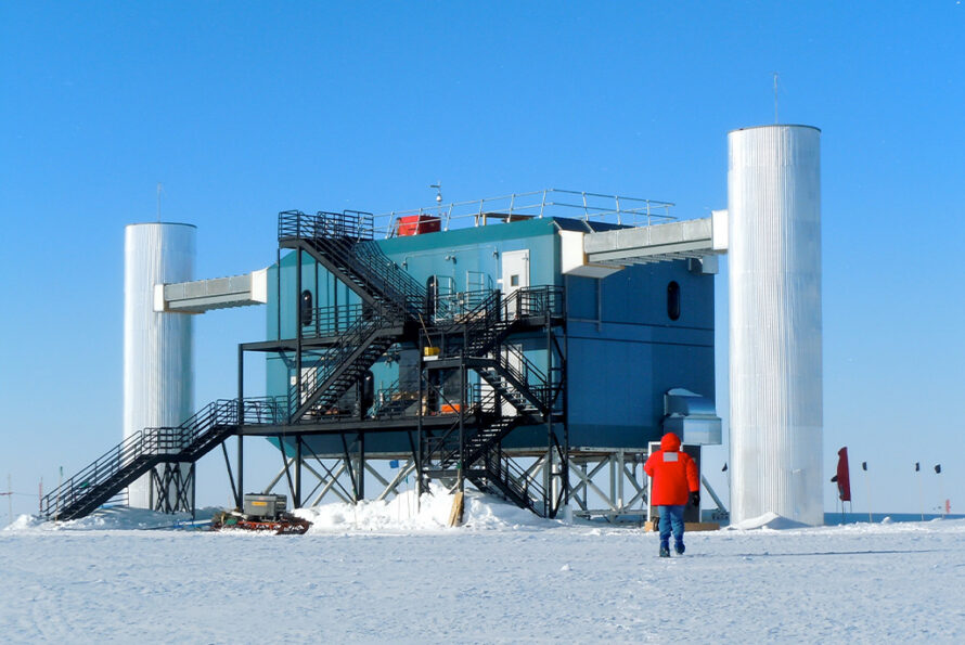 In the snow at the South Pole, a person in a bright red parka walks toward an enclosed, two-story scientific facility with a system of criss-crossing black ladders on the side, and it's sitting on top of suspension girders and suspended between two (2) three-story-tall white towers.